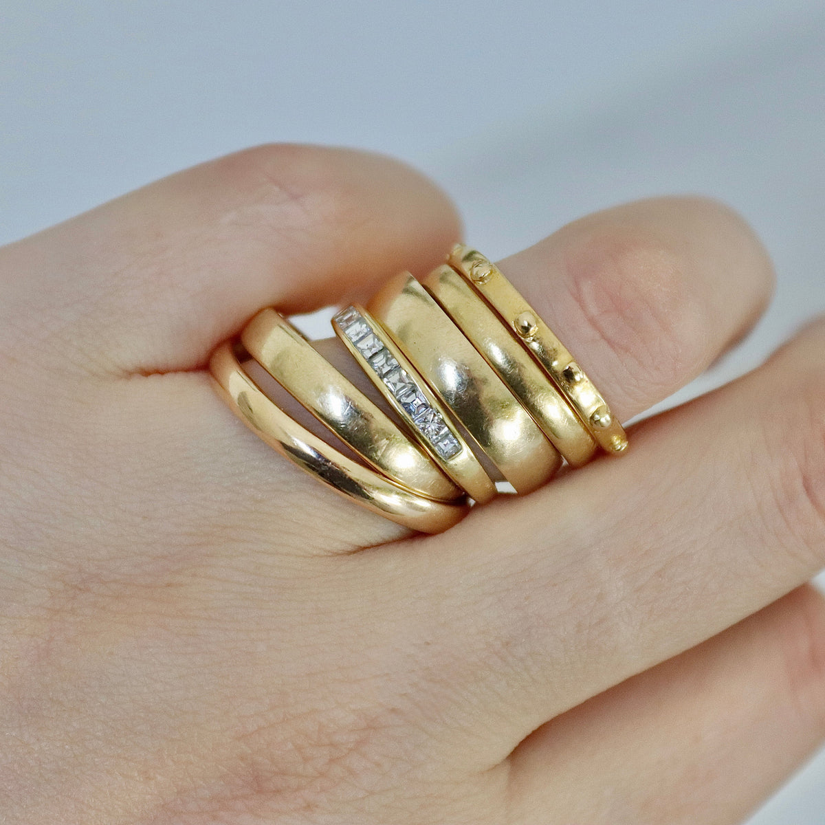 A close-up of a hand wearing a stack of five gold rings, including one with a row of small clear stones, against a soft light gray background.